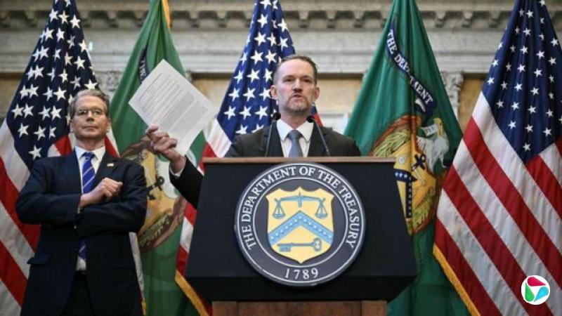 身穿西装的史考特·贝森特站在讲台前的杰米森·格里尔身后,格里尔手持文件面对镜头发言,背后是美国国旗。