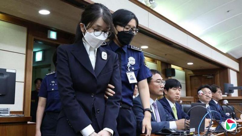 一名身穿黑色套装、戴着口罩与眼镜的女子在一名警官挟持护送下进入法院。她的手臂被警官扶着,而后方桌旁坐着四名身着西装的男子