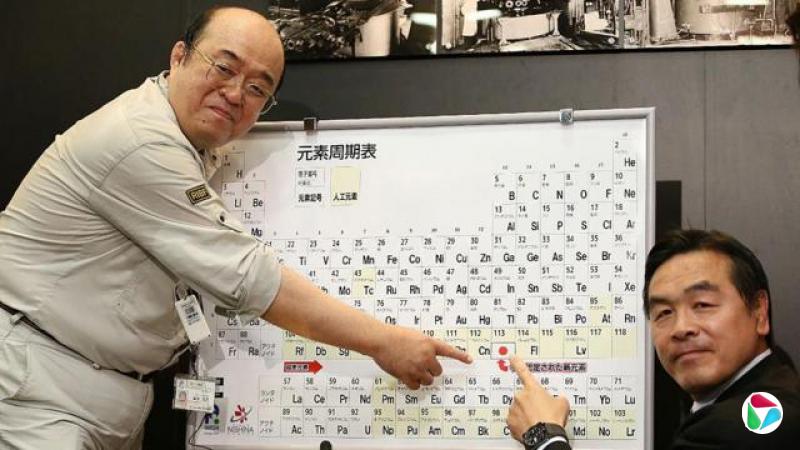一张照片显示两名男子指着一幅装框的元素周期表中的第113号元素,该元素旁标有日本国旗