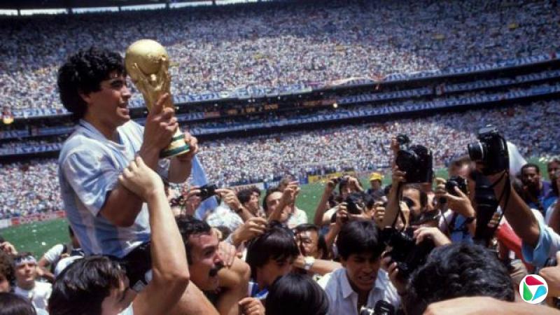 阿根廷队队长迭戈· 马拉多纳(Diego Maradona,马勒当拿)在1986年赛事中夺冠后,被球迷抬在肩上,高举世界杯奖杯庆祝;画面中有人正为他拍照,背景可见看台上成千上万的观众以及足球场一角