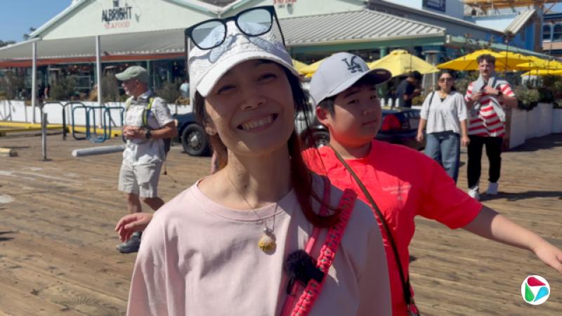 浅井舞子站在画面中央,戴着一顶棒球帽,帽簷上方放着一副眼镜。她的儿子站在她身后,穿着红色上衣。他们站在一条木制步道上,背景是一家海鲜餐厅。