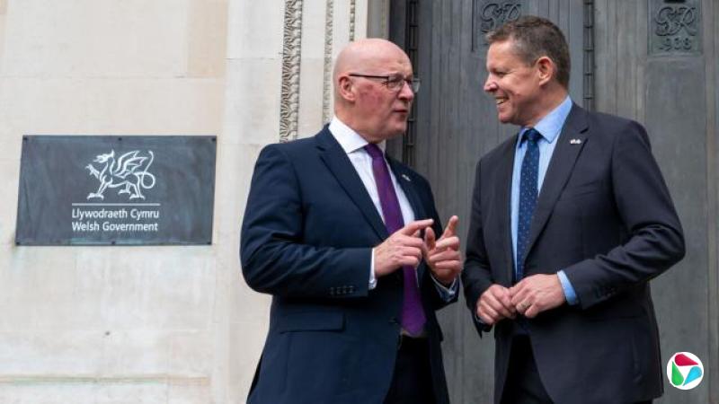 约翰·斯文尼(John Swinney)、杨伟思(Rhun ap Iorwerth)站在威尔士政府总部的一扇金属门外,从照片的左侧可以看到一块铭牌。