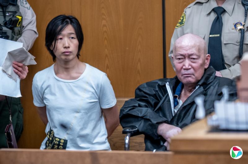 张莎莎身穿白色T恤、双手放在身后,在法庭内坐在宣国军旁边。宣国军是一名秃头的中年男子,两人身后各有执法人员站立。